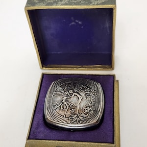 May include: A vintage silver-toned compact mirror with a fairy design, presented in a box with a purple velvet interior. The box has a green exterior with gold trim. The compact is square with a detailed relief of a fairy and floral elements.