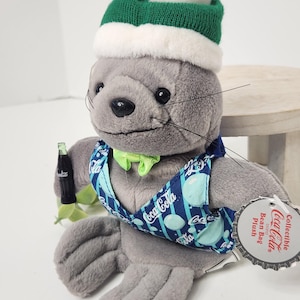 Puede incluir: Un peluche gris con forma de foca que lleva un gorro de punto verde y blanco, un chaleco azul y blanco con logotipos de Coca-Cola y una pajarita verde. La foca sostiene una botella de Coca-Cola en miniatura. Un peluche coleccionable.