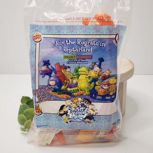 May include: Sealed Burger King toy featuring Rugrats characters in Reptarland. The plastic packaging displays colorful cartoon graphics and the text "Join the Rugrats in Reptarland." The package contains a toy and is not recommended for children under 3 years of age.