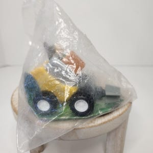 May include: A toy bulldozer in a clear plastic bag. The bulldozer is yellow, blue, and black with white wheels. It sits on a small, light-colored wooden stool.