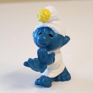 May include: A blue Smurf figurine wearing a white hat and shirt, with a yellow ball on top of the hat. The figurine is in a pose with its hands up, and it has black eyes.