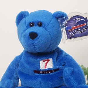 May include: A blue plush teddy bear with a white and red "7" patch on its chest. The bear has black eyes and a black nose. A tag with the Buffalo Bills logo and "D. Flutie" is attached.