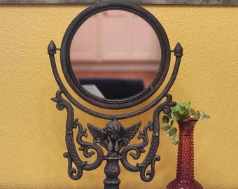 Op de afbeelding: Een decoratieve toilettafelspiegel met een ronde lijst en een sierlijke voet. De spiegel wordt ondersteund door een donker metalen standaard met een engelenfiguur. Een kleine rode vaas met een textuur en groene takjes staat ernaast.