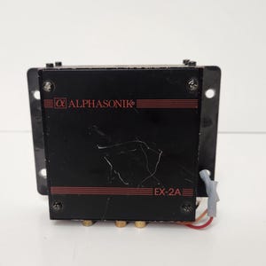 May include: A black Alphasonik EX-2A amplifier with red accents and gold-colored input jacks. The amplifier is mounted on a black metal bracket with mounting holes. The Alphasonik logo is visible on the front.