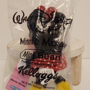 May include: A Minnie Mouse Mini Bean plush toy in a clear plastic bag. The toy is black with a red and white polka dot bow and dress, and yellow shoes. The bag has the Walt Disney World and Kellogg's logos.