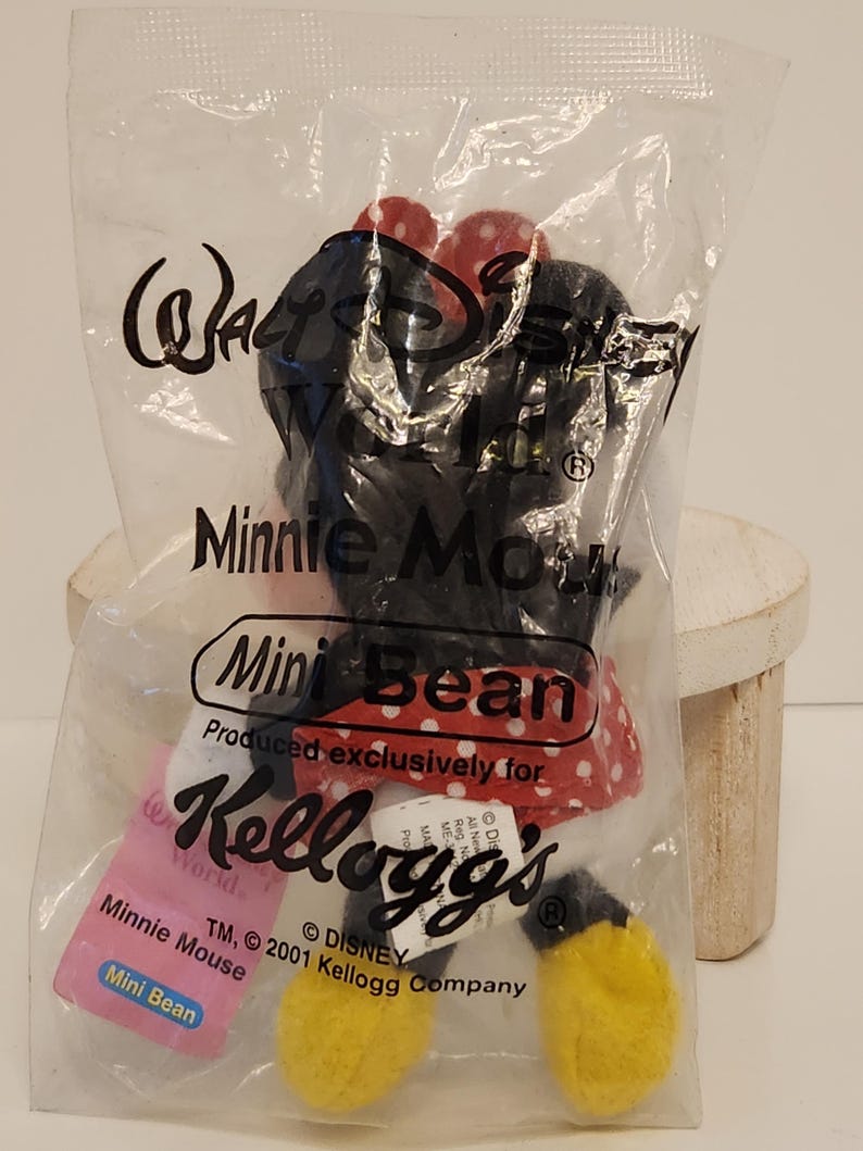 May include: A Minnie Mouse Mini Bean plush toy in a clear plastic bag. The toy is black with a red and white polka dot bow and dress, and yellow shoes. The bag has the Walt Disney World and Kellogg's logos.