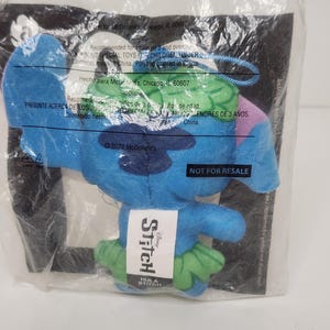May include: A blue plush Stitch toy from the Disney movie, in a clear plastic bag. The toy has green accents and a tag that says "Stitch" and "Hula Stitch". The bag also has text that says "Not for Resale".