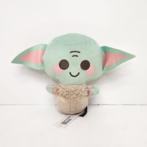 May include: A plush toy of a character with large ears, a green head, and a tan body. The toy has pink accents on the ears and cheeks, with a small tag attached. The character has a smiling expression.