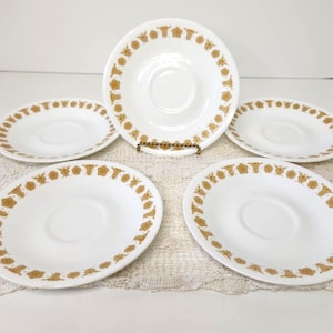May include: A set of five white saucers featuring a circular gold-colored floral pattern along the rim. Each saucer has a central indentation for a cup, with a smooth, glossy finish and a consistent repeating botanical motif in gold.