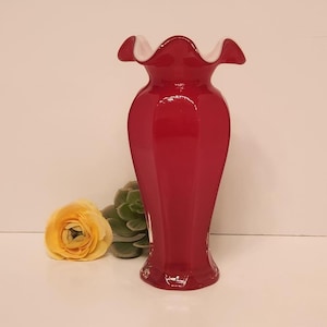 May include: A tall, red glass vase with a ruffled white rim. The vase has a faceted design and is displayed with a yellow flower and a green succulent. The vase is a decorative item.
