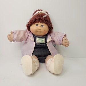 May include: A Cabbage Patch Kids doll with brown yarn hair styled in a ponytail with a white ribbon. The doll wears a light purple zip-up jacket over dark gray denim overalls with a green logo patch, and white socks on its feet.