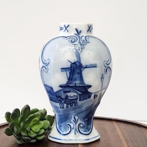 May include: A white ceramic vase featuring a blue hand-painted scene of a windmill and a horse-drawn carriage. The vase has a bulbous shape with ornate blue scrollwork borders and a narrow neck marked with two blue X symbols at the top rim.