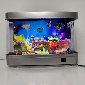 May include: A rectangular, silver-framed light box featuring a Las Vegas-themed display. The illuminated scene includes iconic landmarks, casino chips, dice, and the words "Las Vegas" and "Flamingo". The box has a clear front and a black on/off switch.