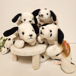 May include: Four plush Dalmatian dog toys. Each toy is white with black spots and black ears. One toy is on a small, round, white wooden table. The toys have black eyes and noses.