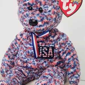 May include: A plush teddy bear with a red, white, and blue speckled pattern. The bear has a black nose and eyes, and a ribbon around its neck. A patch on the chest reads "USA". A heart-shaped tag is attached.