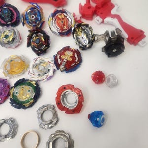 Puede incluir: Una colecci&oacute;n de coloridos juguetes y lanzadores de peonzas. Las peonzas presentan varios dise&ntilde;os con detalles en rojo, azul, dorado y plateado. Tambi&eacute;n se incluyen varios lanzadores rojos y piezas adicionales, todo dispuesto sobre una superficie blanca.