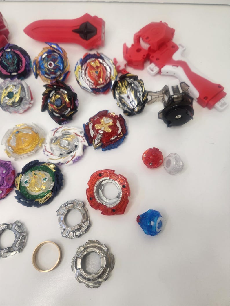 Puede incluir: Una colecci&oacute;n de coloridos juguetes y lanzadores de peonzas. Las peonzas presentan varios dise&ntilde;os con detalles en rojo, azul, dorado y plateado. Tambi&eacute;n se incluyen varios lanzadores rojos y piezas adicionales, todo dispuesto sobre una superficie blanca.