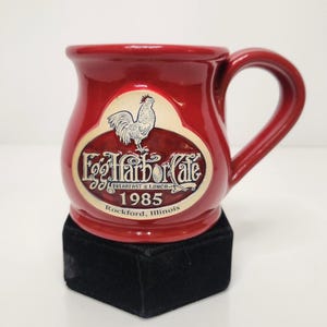 May include: A glossy red ceramic mug featuring a raised cream-colored oval emblem. The emblem displays a rooster illustration, the text Egg Harbor Cafe, Breakfast & Lunch, 1985, and Rockford, Illinois. The mug has a curved body and handle.