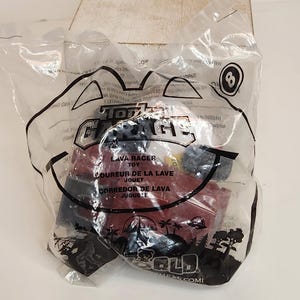 May include: A red and black toy car, labeled "Lava Racer," is sealed in a clear plastic bag. The bag features the text "Total Garage" and the number "8." The packaging includes text in English, French, and Spanish.