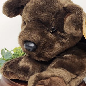May include: A plush brown Labrador Retriever dog toy with a black nose and dark eyes. The stuffed animal is lying down on a wooden base, with its paws extended. The dog's fur is soft and fluffy, and the toy has a realistic appearance.