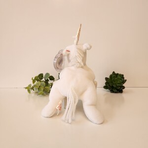 K&ouml;nnte beinhalten: Wei&szlig;es Pl&uuml;sch-Einhorn-Spielzeug mit goldenem Horn und wei&szlig;em Schwanz. Das Spielzeug sitzt auf einer wei&szlig;en Oberfl&auml;che.