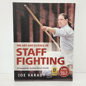 May include: A book titled The Art and Science of Staff Fighting: A Complete Instructional Guide by Joe Varady. The cover features a person in a white martial arts uniform holding a wooden staff, with a red badge stating 9 Levels Easy to Expert.