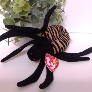 May include: A black plush spider toy with a striped brown and tan body. The spider has eight long, black legs and red eyes. A red heart-shaped tag with a yellow star is attached to the spider.