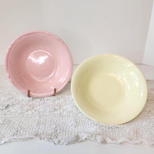 May include: Two ceramic bowls, one light pink and the other pale yellow, are displayed on a white lace doily. The bowls have a simple, classic design with a slightly raised rim. The pink bowl is on a gold-colored stand.