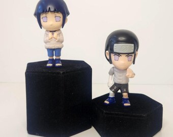 May include: Two small anime-style figurines featuring characters with dark hair and purple eyes. One figure wears a light-colored hooded jacket with blue shorts, while the other wears a grey shirt, black shorts, and a headband with a spiral symbol.