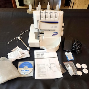 May include: A white and cream-colored Juki MO-1000 serger sewing machine with accessories. The machine has a curved design and a blue and gold accent. Included are a foot pedal, needles, a DVD, and a manual.