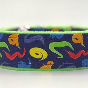May include: A dog collar with a dark blue fabric featuring colourful worm illustrations in yellow, orange, red, green, and purple. The collar has a bright green trim.