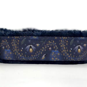 May include: A navy blue dog collar with a pattern of glowing fireflies and leaves. The collar has a soft, dark blue faux fur lining and a silver metal D-ring for leash attachment.