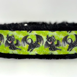 May include: A dog collar featuring a repeating pattern of a black dragon on a lime green background. The collar is trimmed with black, fluffy material. The dragon has its tongue sticking out.