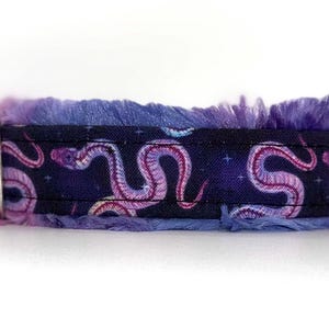 May include: A dog collar featuring a dark blue fabric with a repeating pattern of pink snakes and stars. The collar has a fluffy, purple trim and a silver metal loop for attaching a leash.