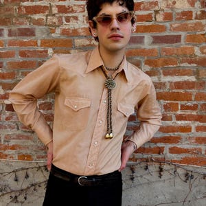 May include: A tan button-down shirt with two chest pockets and a bolo tie with a decorative pendant and gold-colored tips. The shirt is paired with black pants and a black belt with a silver buckle. The background is a brick wall.