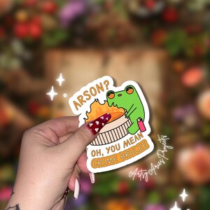 Könnte beinhalten: Aufkleber mit einem grünen Frosch, der eine rosa Sprühflasche hält, mit einem brennenden Crème brûlée-Dessert. Der Text auf dem Aufkleber lautet "ARSON? OH, YOU MEAN CRIME BRULEE."