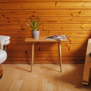 May include: A white armchair with a wooden base sits next to a small wooden table with a potted plant and a magazine on top. A blue rocking chair with a wooden frame is in the background. The chairs and table are in front of a wood panelled wall.