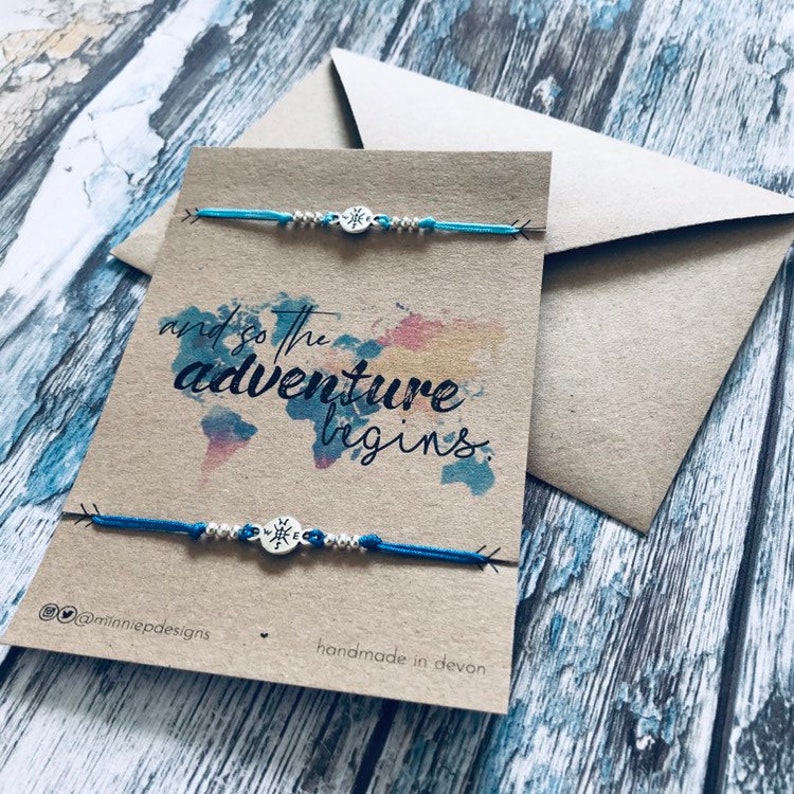 May include: Two blue friendship bracelets with silver compass charms and white beads. The bracelets are displayed on a card with the text "and so the adventure begins" and a watercolour world map. The card also includes the text "@minniepdesigns" and "handmade in devon".