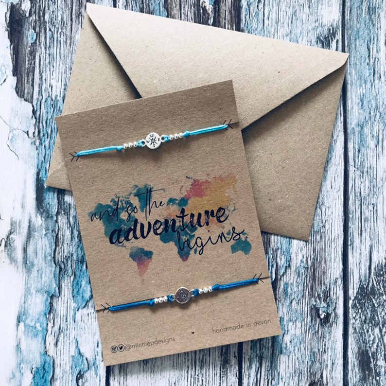May include: Two blue friendship bracelets with silver compass charms on a brown card with the text "and so the adventure begins" and a watercolour world map. The card is handmade in Devon.