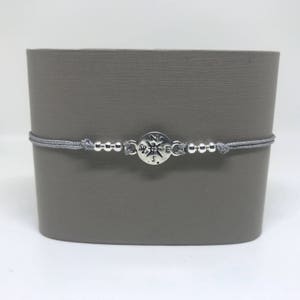 May include: A grey cord bracelet with a silver compass charm. The compass charm has the letters N, S, E, and W engraved on it.