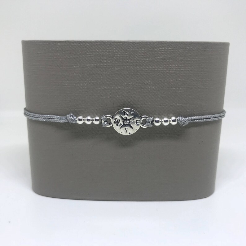 May include: A grey cord bracelet with a silver compass charm. The compass charm has the letters N, S, E, and W engraved on it.