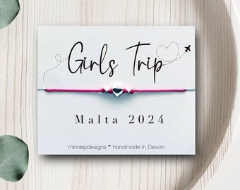 Può includere: Un braccialetto di corda rosa con un ciondolo a forma di cuore argentato e il testo "Girls Trip Malta 2024" su un cartoncino bianco. Il cartoncino riporta anche "minniepdesigns handmade in Devon".