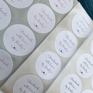 Może przedstawiać: Arkusz białych okrągłych naklejek z tekstem "Handmade with love by Kirsty" w fioletowym piśmie odręcznym. Naklejki są ułożone w rzędach i kolumnach.