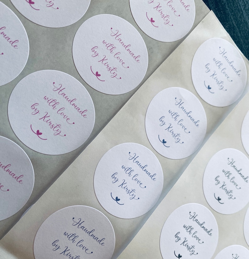 Może przedstawiać: Arkusz białych okrągłych naklejek z tekstem "Handmade with love by Kirsty" w fioletowym piśmie odręcznym. Naklejki są ułożone w rzędach i kolumnach.