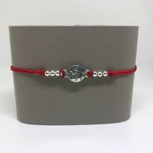 May include: Red string bracelet with silver beads and a compass charm. The compass charm has the letters N, S, E, and W engraved on it.