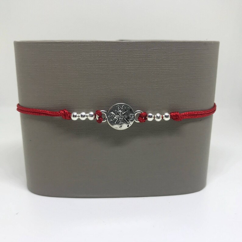 May include: Red string bracelet with silver beads and a compass charm. The compass charm has the letters N, S, E, and W engraved on it.