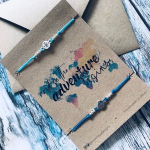 May include: Two blue friendship bracelets with silver compass charms on a brown card with a watercolour world map and the text "and so the adventure begins".
