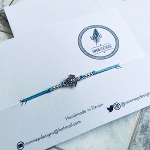 May include: A light blue bracelet with a silver turtle charm and silver beads. The bracelet is handmade in Devon and the text "minniepdesigns@hotmail.com" and "@minniepdesigns" are printed on the card.
