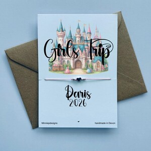 May include: A thin string bracelet with a small black heart charm attached to a white display card. The card features a colourful illustration of a castle, the text Girls Trip, Paris, and 2026, along with minniepdesigns and handmade in Devon.