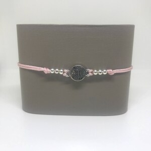May include: A pink string bracelet with silver beads and a silver compass charm. The compass charm has the letters N, S, E, and W engraved on it.